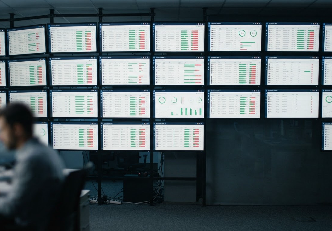 Parede de monitores exibindo dezenas de interfaces de aplicativos web em avaliação, com marcadores de testes aprovados e reprovados comparando desempenho individual e em equipe