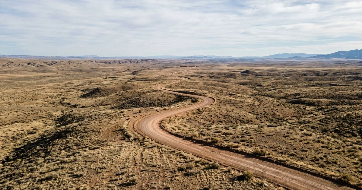 Vista aérea de terreno árido e remoto nos Estados Unidos, com estrada de terra sinuosa cortando a paisagem seca e horizonte amplo sem construções
