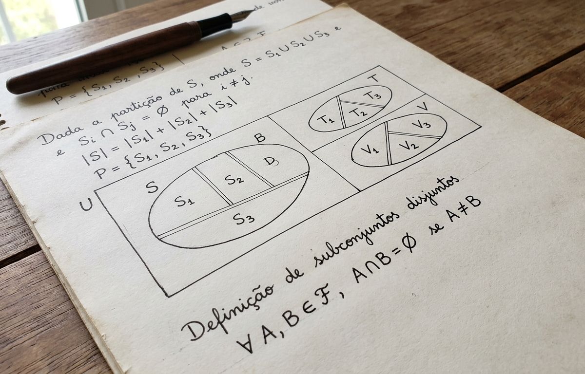 Manuscrito matemático sobre mesa com diagramas de conjuntos divididos em subconjuntos disjuntos por linhas finas, símbolos de teoria dos conjuntos escritos à mão e caneta apoiada, iluminados por luz lateral suave